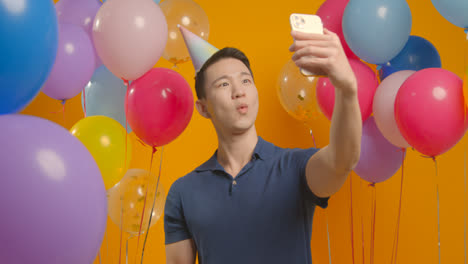 Studioporträt-Eines-Mannes,-Der-Ein-Selfie-Mit-Partyhut-Macht-Und-Geburtstag-Feiert,-Umgeben-Von-Luftballons-1